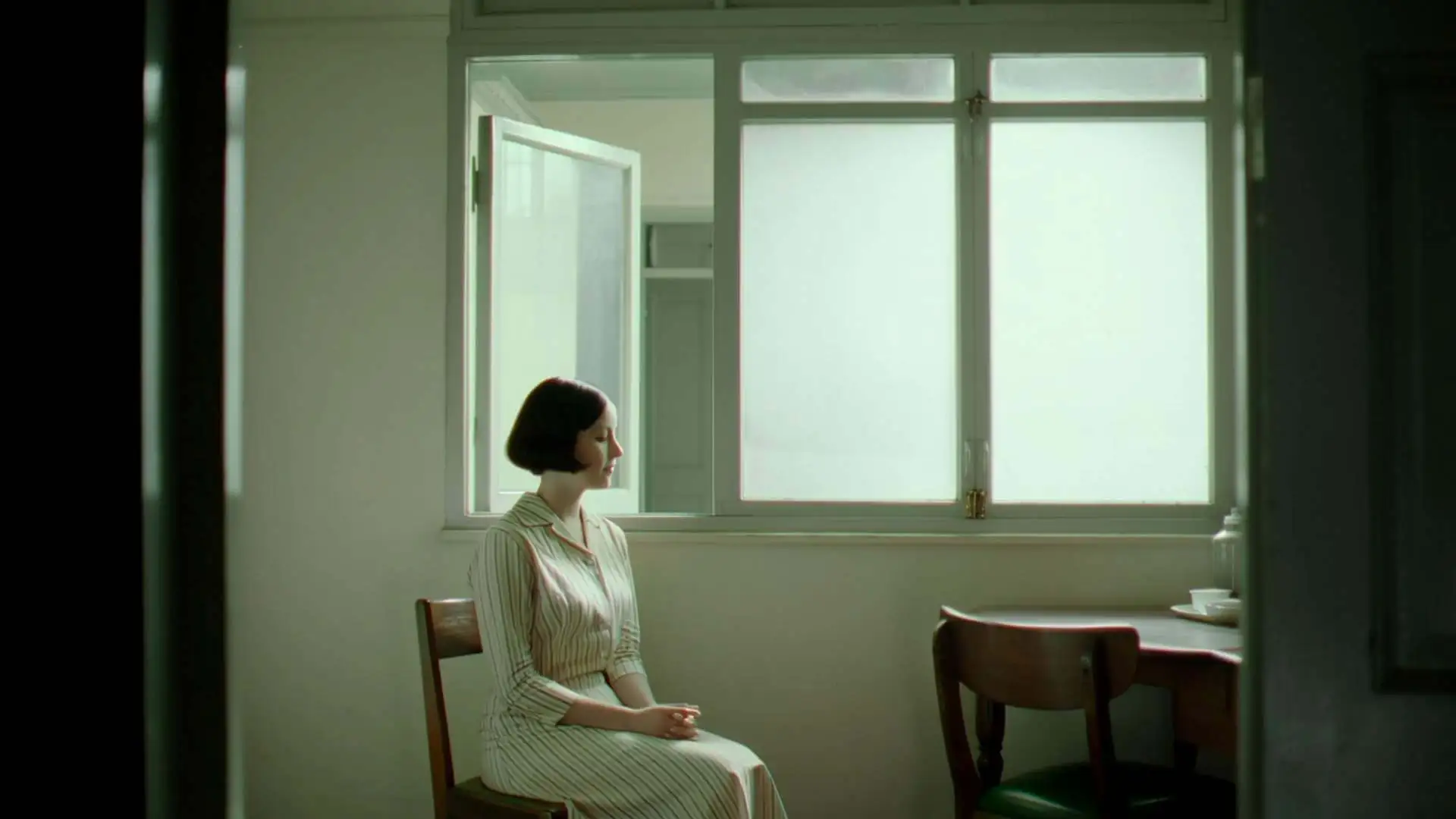 A woman in a striped dress sits quietly on a wooden chair beside an open interior window. A man from the adjoining room slowly leans through and gives her a brief, tender kiss, then gently pulls back. Fixed medium-wide shot, camera static throughout. Soft natural daylight from a background window, faded greenish film tone, bright window bloom. Late-1970s 35mm arthouse style transferred to worn VHS — visible grain, gate weave, chromatic aberration, dust specks, restrained quiet intimacy.