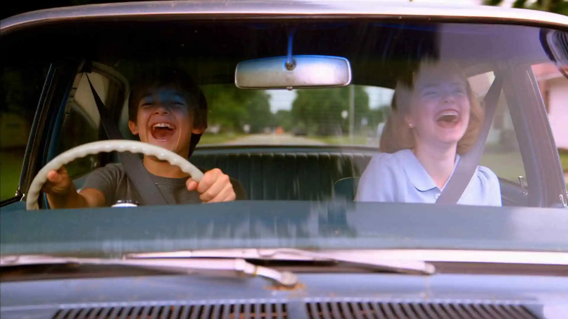 Two teenagers — a boy driving, a girl in the passenger seat — burst into uncontrollable laughter together inside a small moving car on a quiet suburban road. Hood-mounted camera looks inward through the windshield, steering wheel visible in foreground. Tree reflections slide across the glass, soft late-afternoon light and shadow cross their faces. Camera shakes slightly from road vibration.