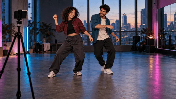 Dos personas adultas bailan frente a un móvil con trípode en un estudio iluminado en azul y magenta.